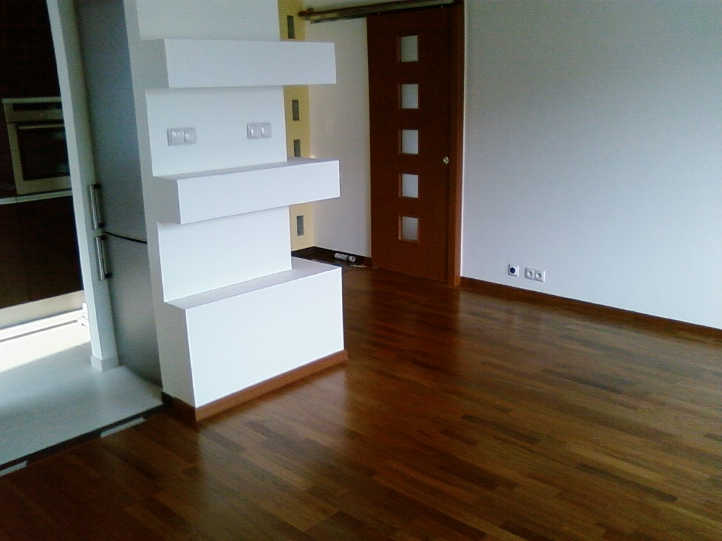 Ciemny, lakierowany parkiet ułożony w jodełkę, z widoczną białą, modernistyczną ścianką działową z półkami i brązowymi drzwiami w tle, oświetlony naturalnym światłem.